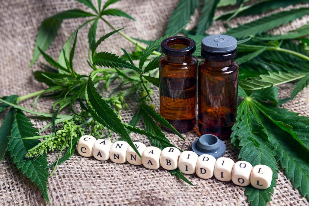 Is CBD safe and what are cbd oil side effects? Image shows two bottles of CBD, hemp leaves, and some letter blocks spelling out cannabidiol.