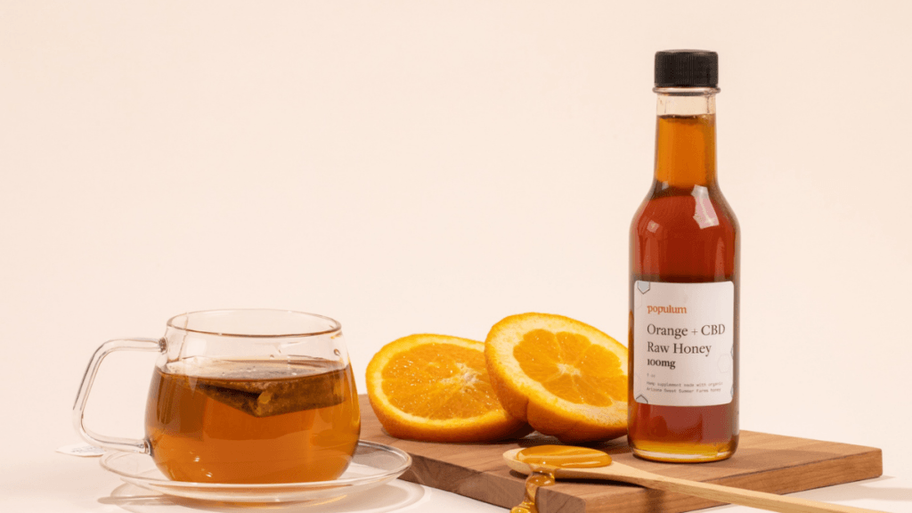 A photo of Populum Orange + CBD Raw Honey posed with a glass mug of tea with teabag, two orang slices, an da spoonful of honey spilling onto the cutting board next to the bottle.