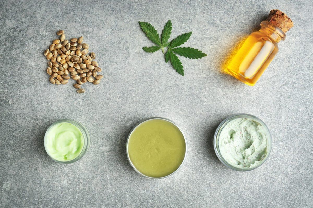 A variety of topical CBD products seen with some hemp seeds, a hemp leaf, and a vial of essential oil. 