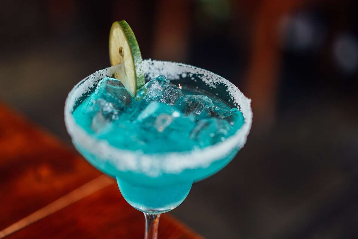 Some people report that CBD cocktails reduce the negative effects of alcohol intoxication. A blue cocktail sits in a cocktail glass with a salted rim and a cucumber garnish.