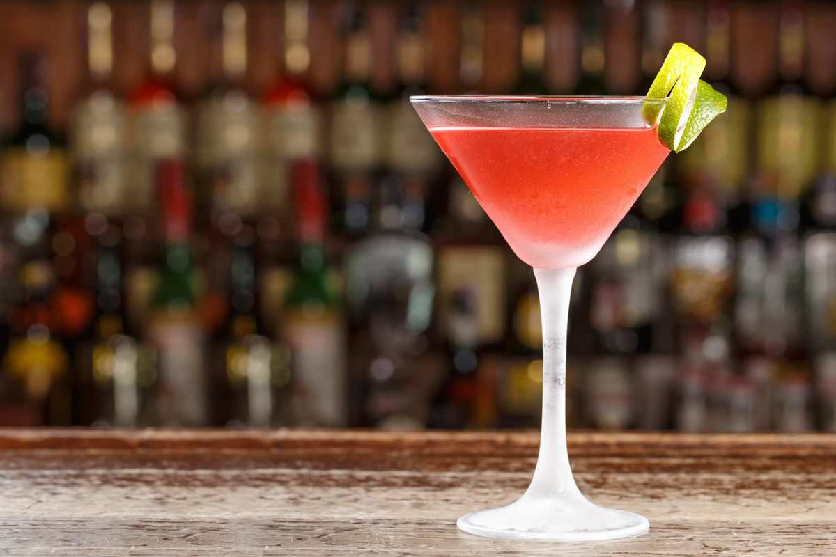 You can start with our CBD-infused Cosmo recipe, but don't be afraid to experiment. The potential of CBD cocktails is nearly endless. A red cocktail in a martini glass, garnished with a lemon peel, sits on a fancy bar with shelves of alcohol in the background.