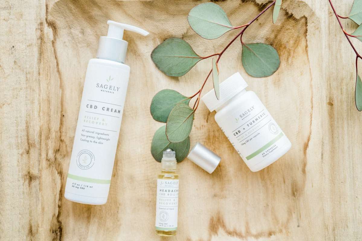 Sagely Naturals Relief and Recovery collection includes a topical CBD cream, a headache roll-on, and CBD capsules with turmeric. In this photo, the products are shot on a wooden background with a sprig of leaves for decoration.