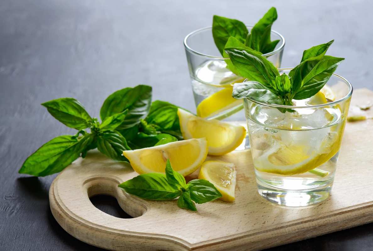 CBD drinks can help you unwind after a long day. A cutting board with lemons, basil and two lowball glasses of basil lemonade.
