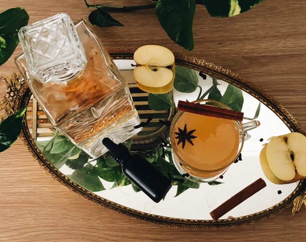 A decanter of bourbon sits on a platter with a bottle of tincture, apples, cloves, and a glass of cider decorated with a cinnamon stick. Chron Vivant's CBD Mulled Apple Cider will warm you up and help relieve the stress of the holidays.