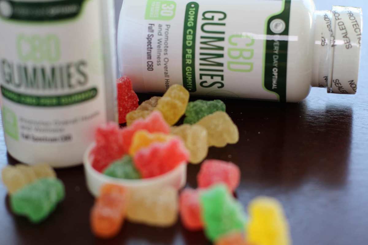 A pile of Every Day Optimal CBD gummies spill from gummy bottle, while another bottle of gummies lies nearby. the cap of a Every Day Optimal CBD gummies, available in 25mg strength, pack potent CBD relief into a delicious sweet & sour candy.