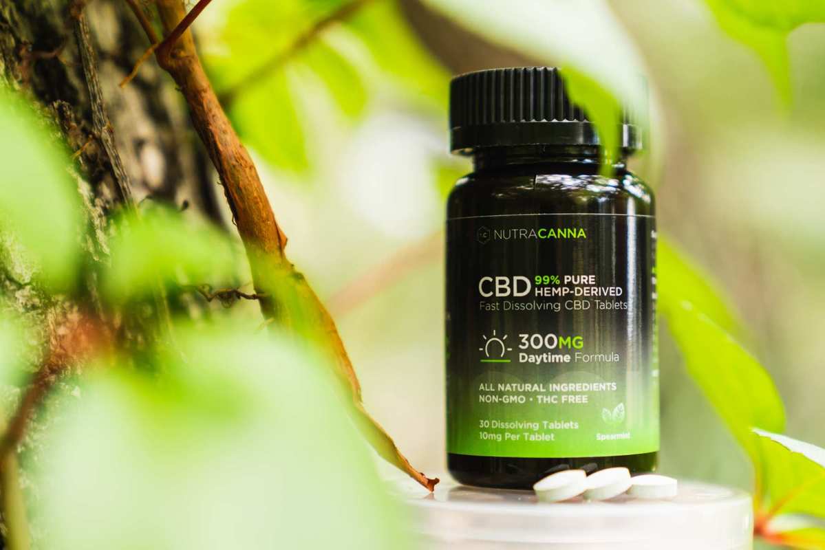 A bottle of NutraCanna Dissolvable CBD tablets posted in a forest, with two of the white round tablets. We were excited to try NutraCanna Dissolvable CBD tablets and the results were rewarding.
