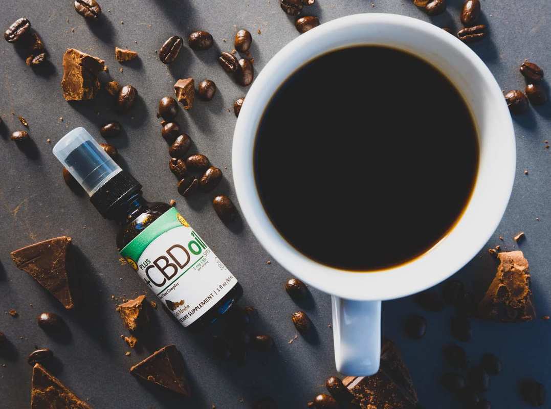 Bottles of PlusCBD Cafe Mocha Spray sits near a cup of black coffee and scattered coffee beans and chocolate chunks. PlusCBD Cafe Mocha Spray helped our review team start our day with a relaxing and flavorful cup.