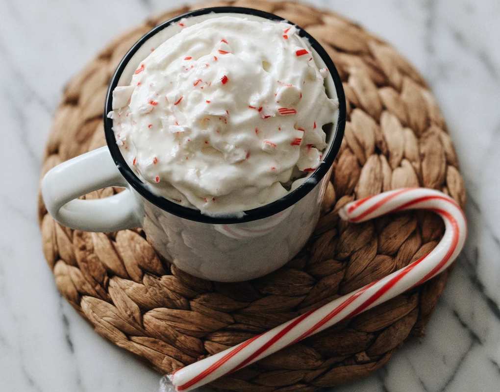 A mug of CBD hot cocoa garnished with whipped cream and candy cane pieces. The mug sits on a woven coaster with another candy cane.