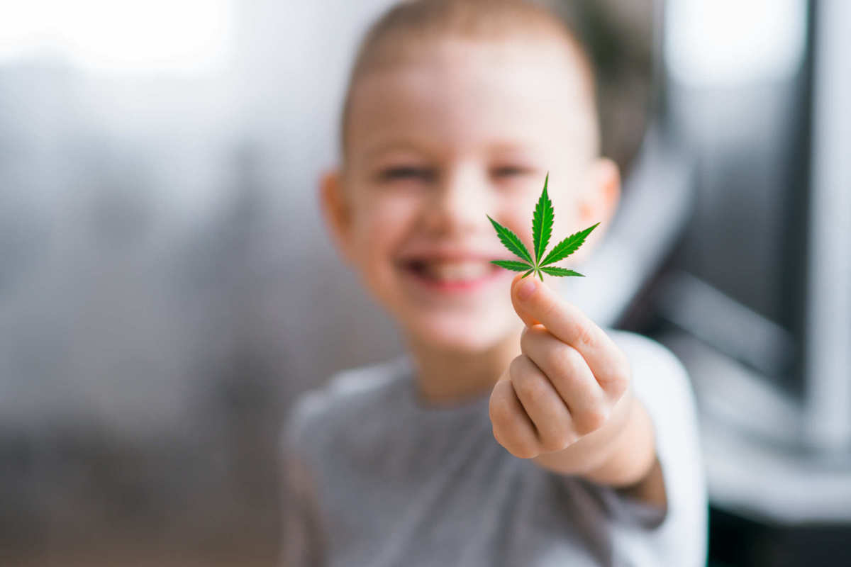 A young child with short hair grins while holding a hemp leaf toward the camera. The British Hemp Association believes that hemp can have a massive positive impact on people in the UK and the planet as a whole, if that government will get out of their way.