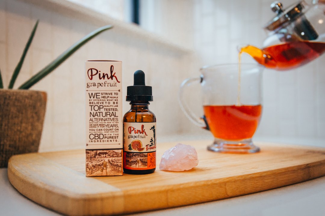 A bottle of Savage CBD Pink Grapefruit CBD Tincture, with accompanying box, sits on a cutting board while a cup of tea is poured into an nearby mug. With its fresh fruit taste, Savage CBD Pink Grapefruit Tincture makes a great addition to tea and CBD cocktails.