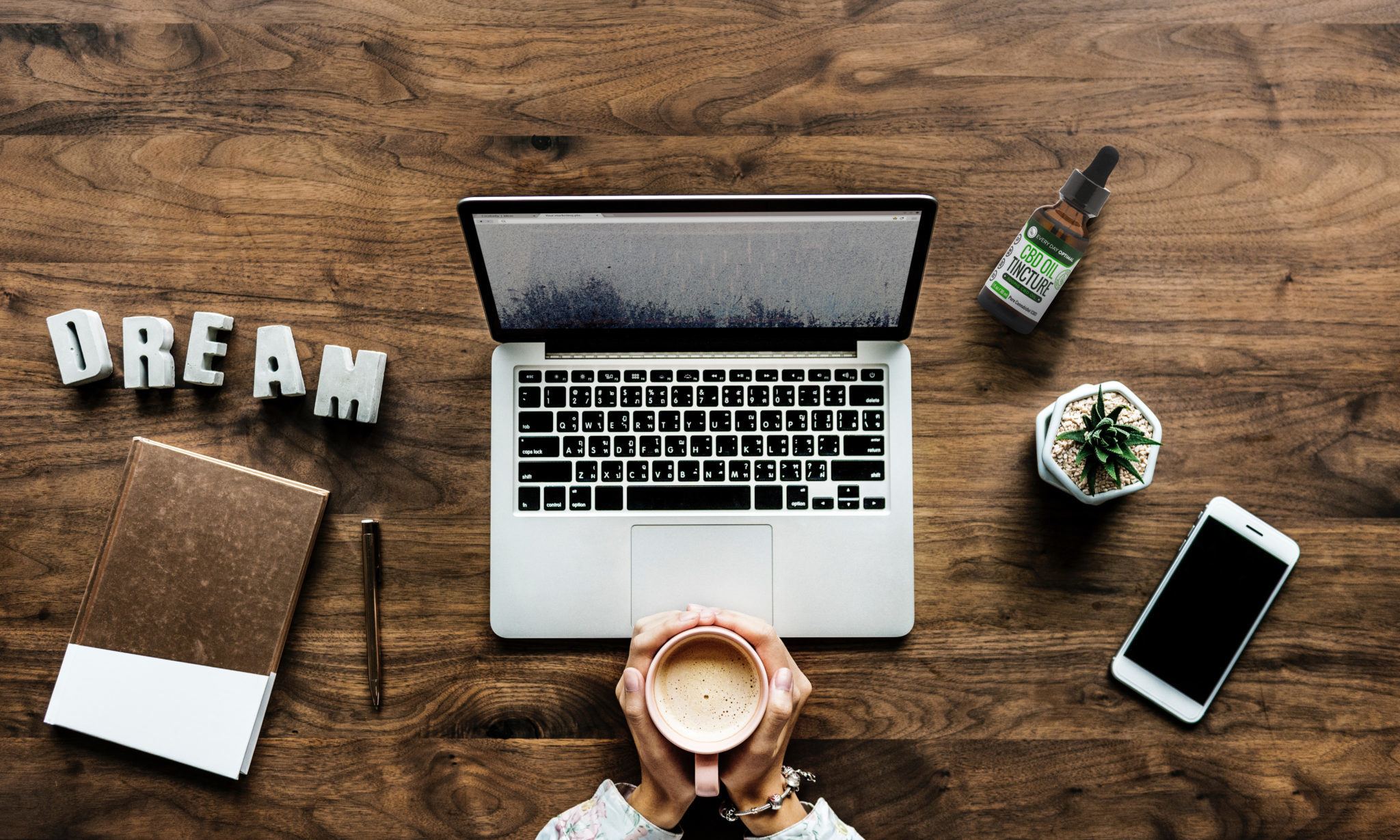 In this article, we compare the benefits of CBD vs CBN and explain how both can help us live better lives. A seated person cups a mug of coffee in their hands in front of a laptop. Arranged nearby are a small plant, a notepad, a smartphone, a bottle of Every Day Optimal CBD, and decorate letters spelling the word DREAM.