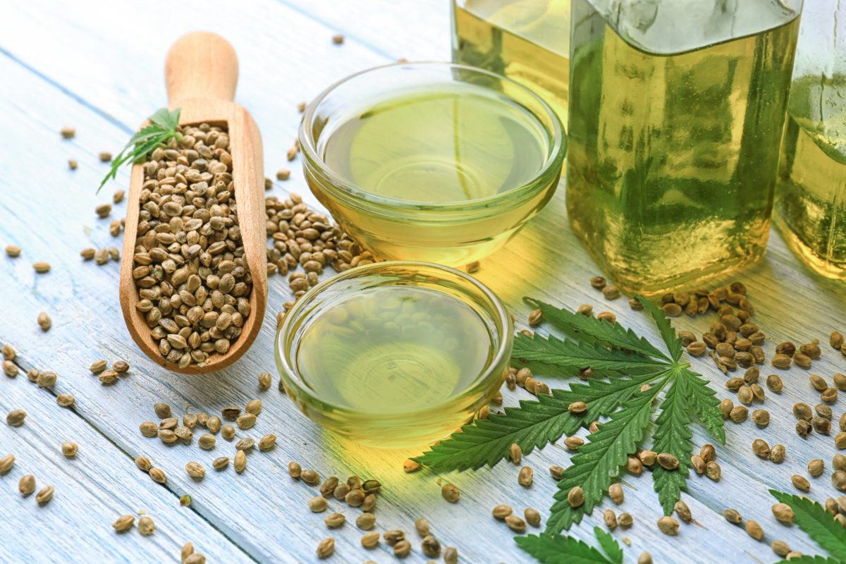 An overflowing scoop of hemp seeds sits near a bowl of hemp seed oil and two bottles of vegetable oil, along with a hemp leaf. CBD tinctures typically combine hemp extract with a carrier oil such as hemp seed oil, MCT oil, or even olive oil.