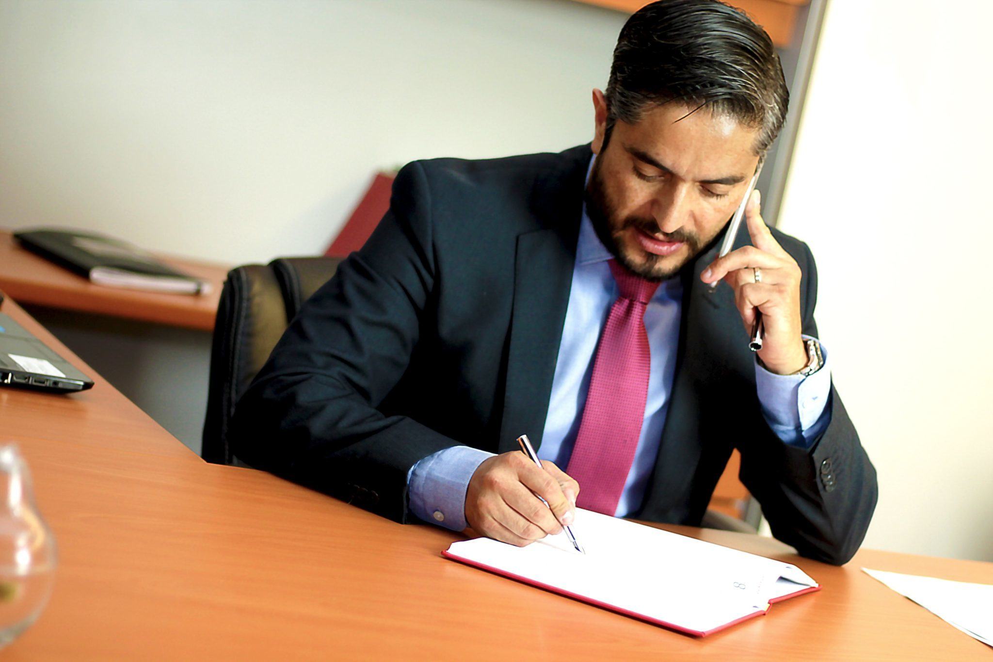Other factors that increase the cost of CBD include retaining a lawyer, hiring an in-house compliance officer, and the complicated nature of banking and insurance in the hemp and cannabis industries. Photo: A lawyer in a suit and tie writes on a notepad while talking on a smartphone in an office.