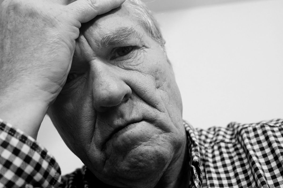 Photo: An older man in a checked button down shirt holds his forehead in despair.