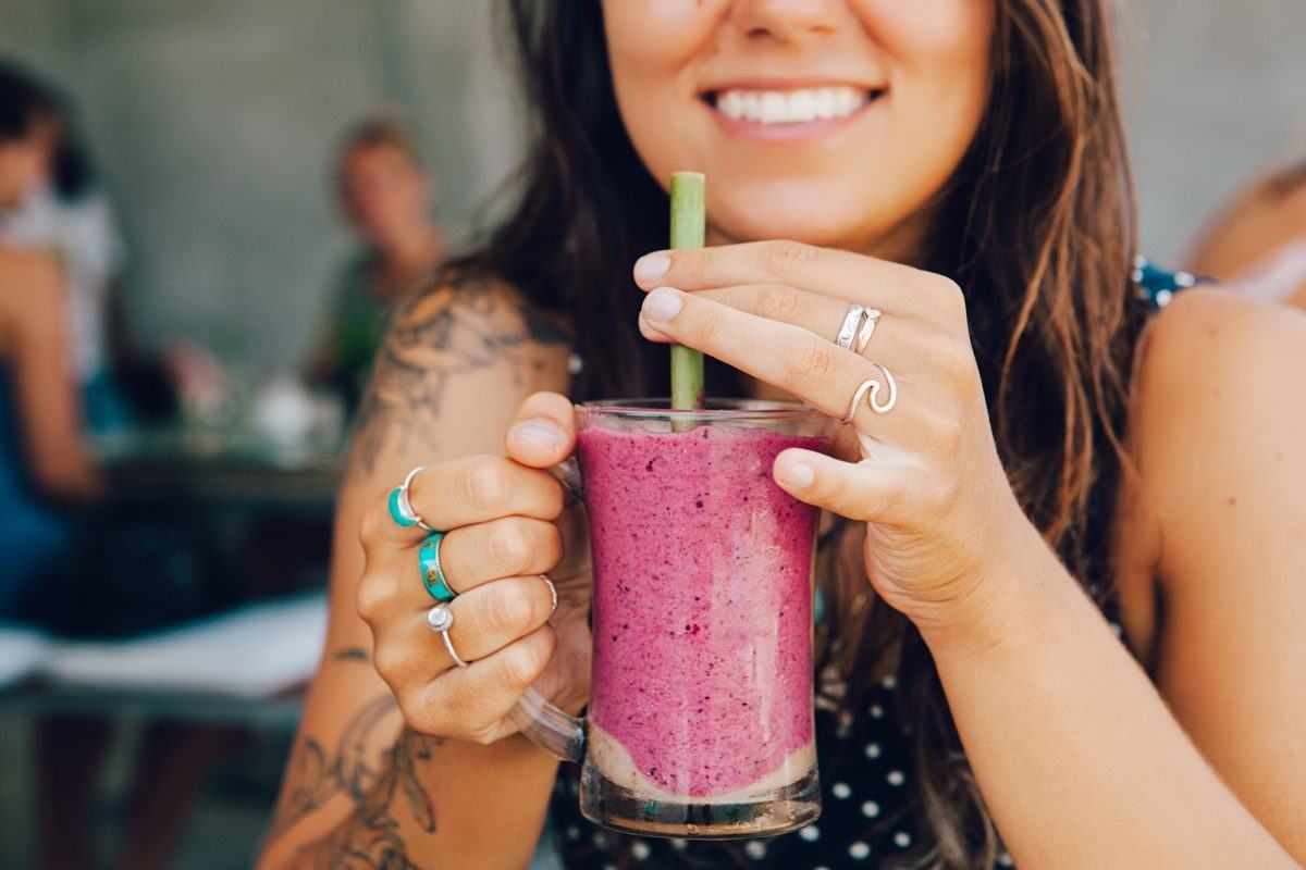 A smiling woman enjoys a easy hemp protein smoothie from a glass mug, with a straw.