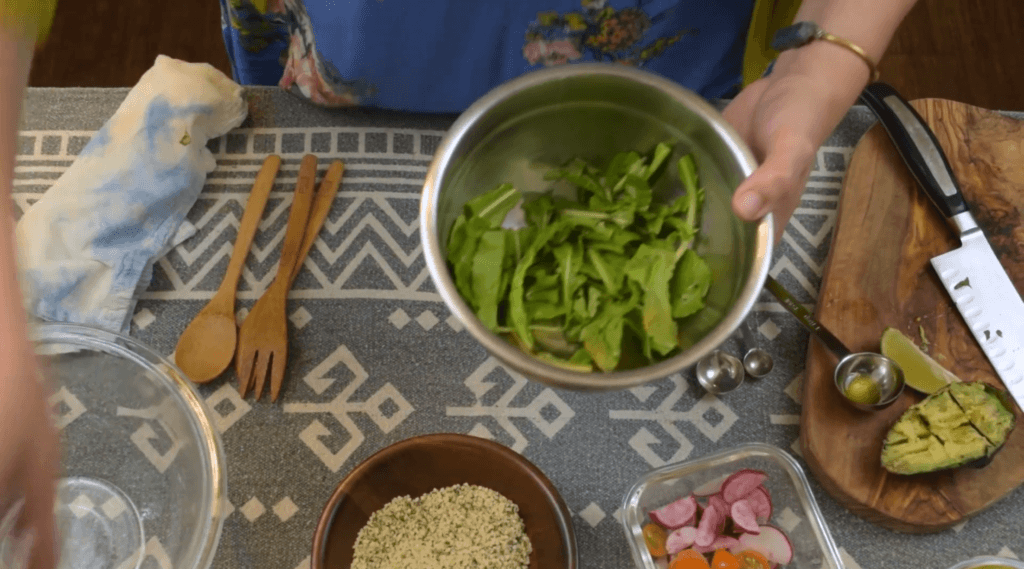 Video screenshot shows Drew assembling the hemp seed salad, with arugula in her bowl, and hemp hearts, strawberries, avocado and other ingredients ready nearby.