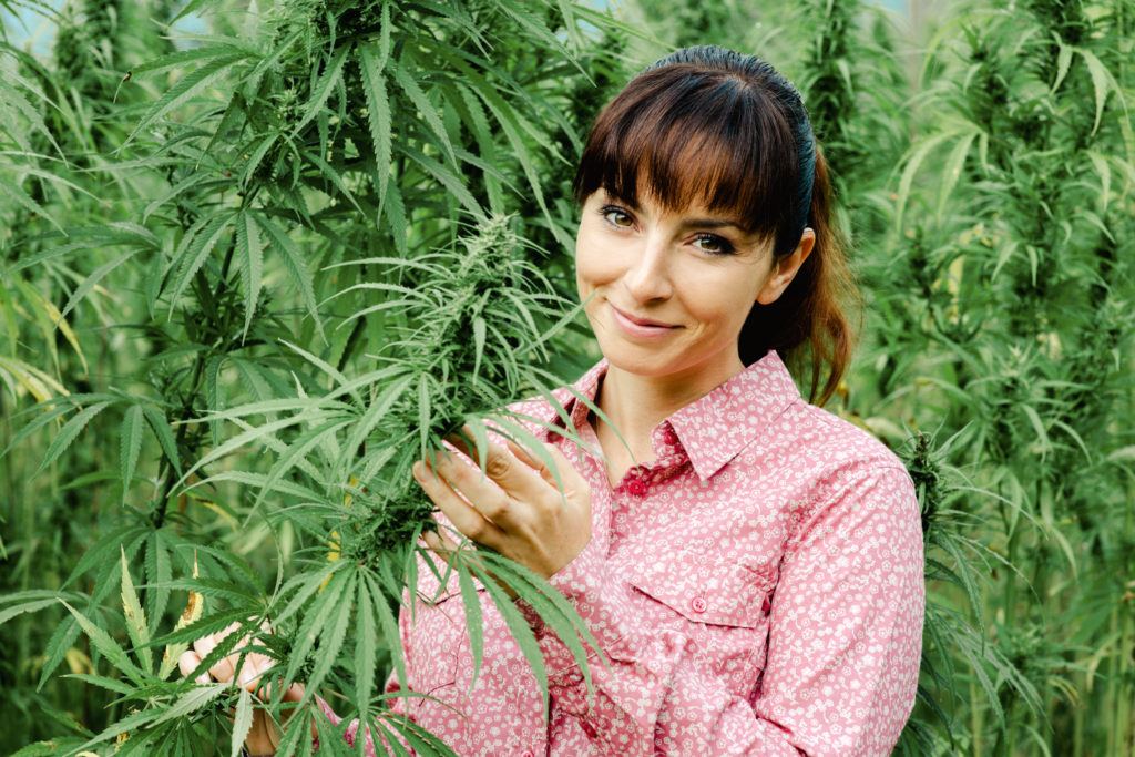 On the Ministry of Hemp Podcast, we're beginning a series on the women of hemp. Photo: A smiling young woman stands in a hemp field, wearing a pink button down. One hand holds part of a leafy hemp plant.