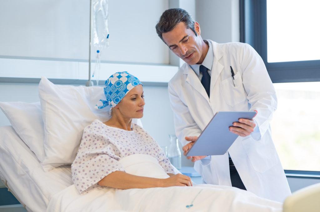 While rare, CBD can interfere with a few medications. It's always important to consult with a medical professional before adding CBD to your cancer treatment. Photo: A doctor consults with a woman in a hospital bed, holding a clipboard. She has a hair wrap on due to cancer treatment hair loss.
