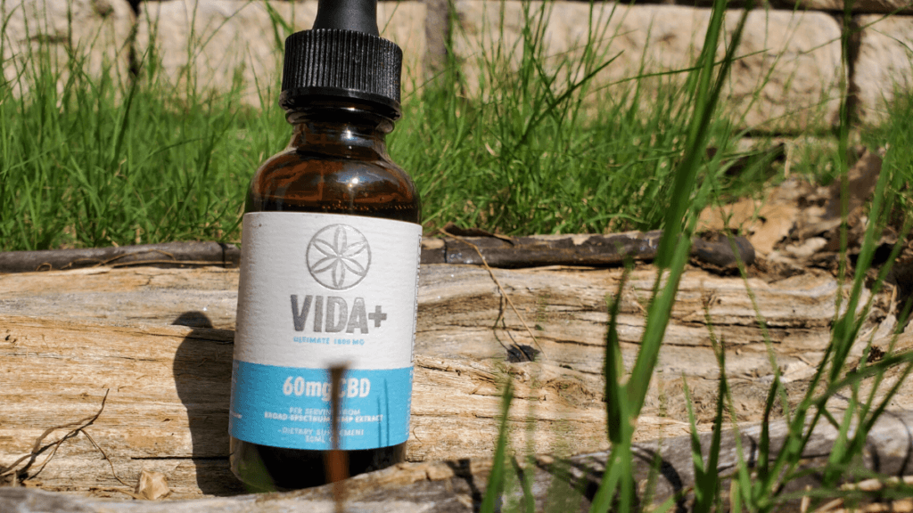 Photo: A bottle of Vida+ Ultimate CBD Oil Tincture posed outside on a wooden log among grass, with a stone wall in the background.