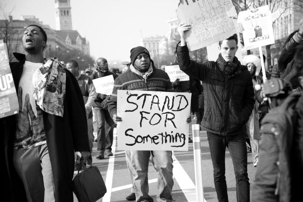 In a black and white photo at a Black Lives Matter protest, a black marcher holds a handwritten sign reading "Stand For Something."