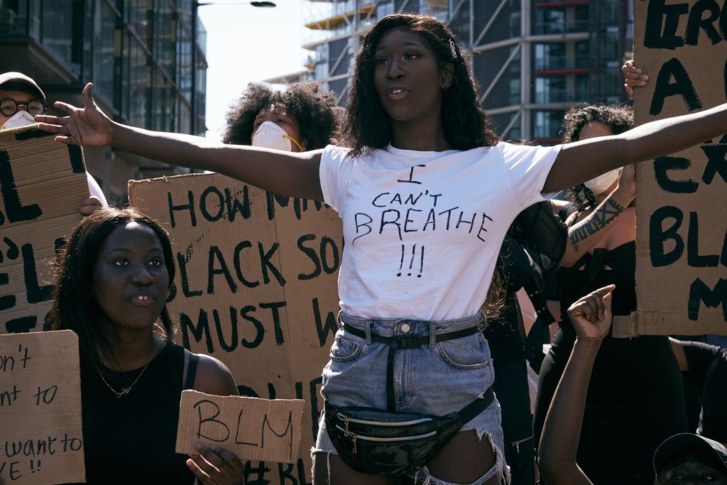 Photo: A Black woman in a handmade "I Can't Breathe" shirt holds her arms open in front of a crowd at a protest. 