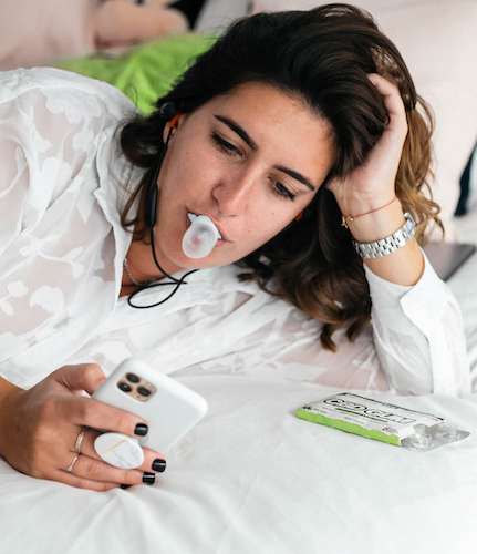 Photo: A white woman with dark brown hair looks at her smartphone while blowing chewing bubble gum in bed. A box of Every Day Optimal CBD gum rests on the bed.