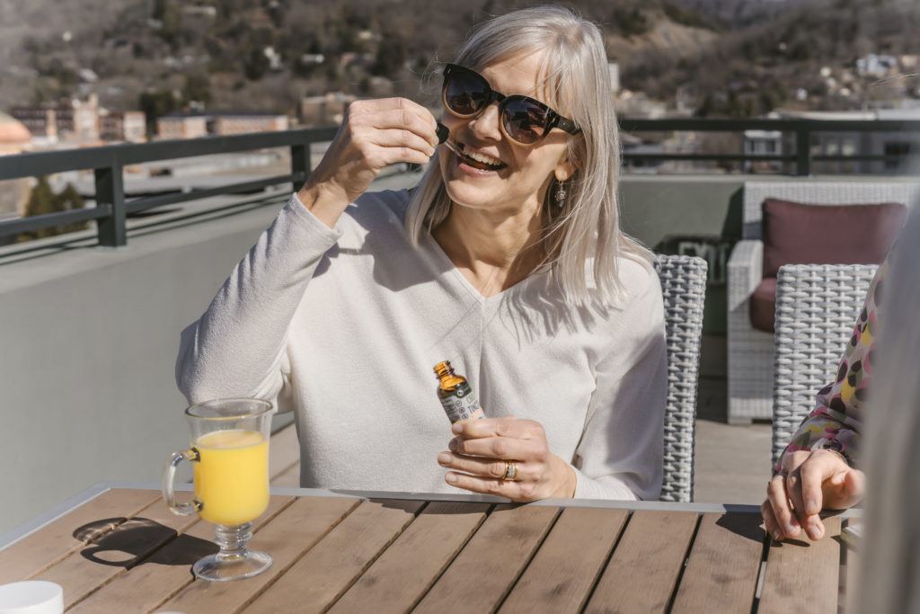 Photo: An older white woman with gray hair smiles as she takes Every Day Optimal CBD oil while sitting at an outdoor cafe.