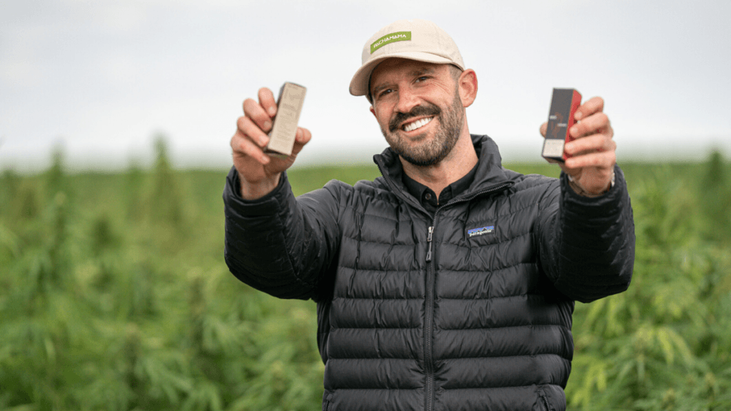 On the Ministry of Hemp Podcast, we learned about how Brandon Stump used CBD for addiction recovery, leading him to found his own company, Pachamama. Photo: Brandon Stump, founder of Pachamama CBD, holds two boxes of his products in a hemp field.