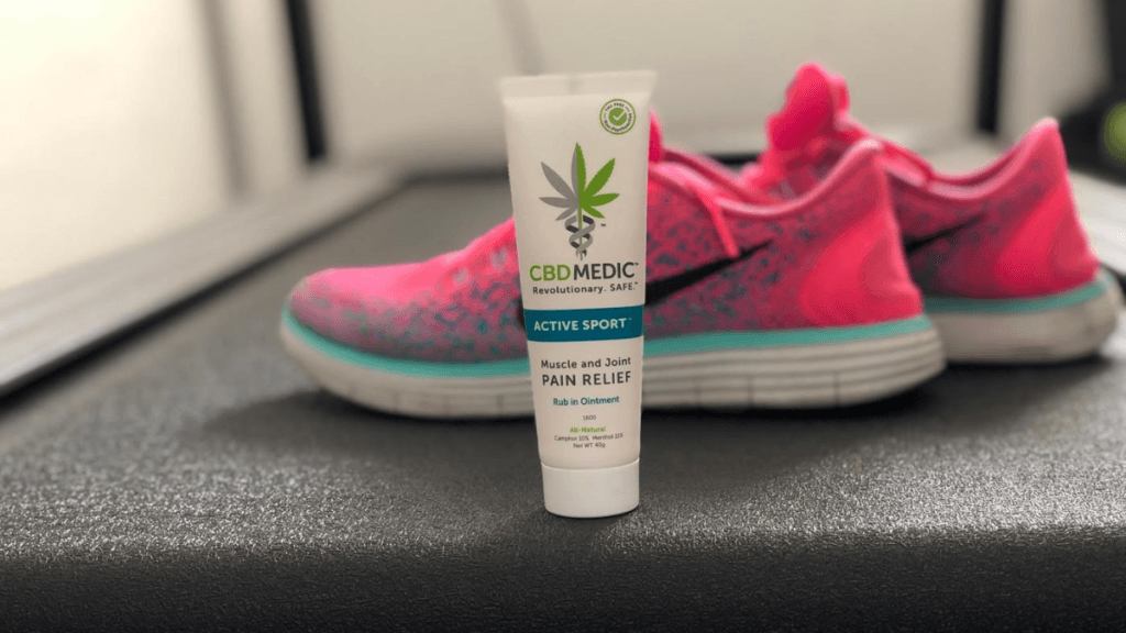 A tube of CBDMEDIC Active Sport Ointment posed with a pair of pink sneakers in a locker room.
