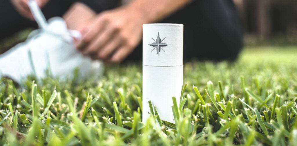 A bottle of Canvas 1839 CBD oil sits on a fake grass surface while, in the background, someone ties their athletic shoes.