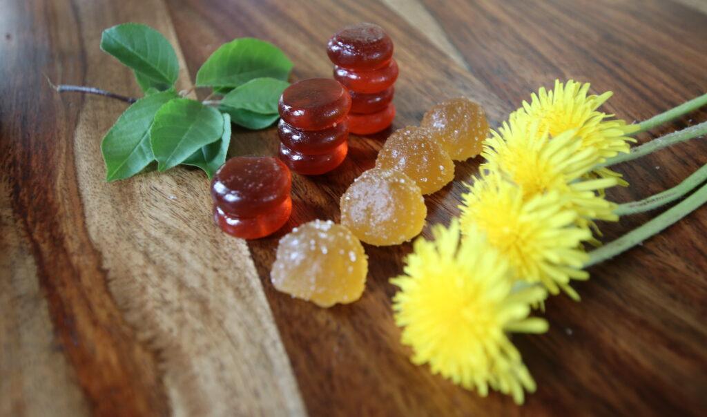 Different styles of CBD gummies or chews decoratively posed with mint leaves and dandelion flower on a richly grained, smooth wooden surface.