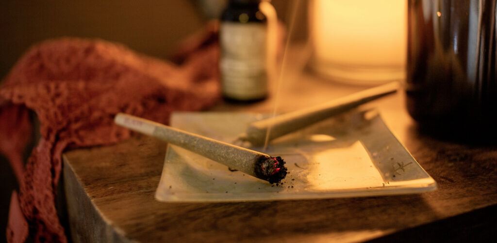 A pre-rolled hemp flower joint smokes in a white ashtray, with another unlit nearby. The ashtray sits on an end table with CBD oil, a tea cup and some fabric in the background.