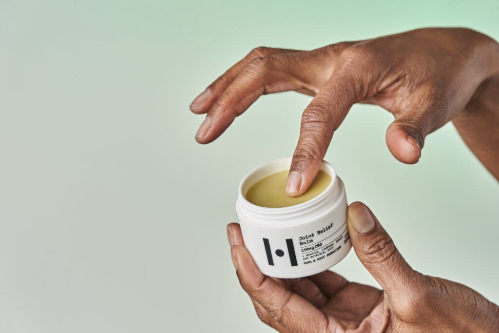 Someone holds the open bottle of Healist Naturals Joint Relief Balml in their hands as they gather some onto a finger for application to sore muscles.