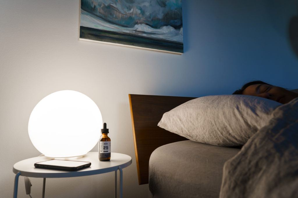 A person sleeps soundly in their bed, with a bottle of Receptra Naturals Serious Rest Tincture on the bedside table near a globe-shaped lamp and a smartphone.