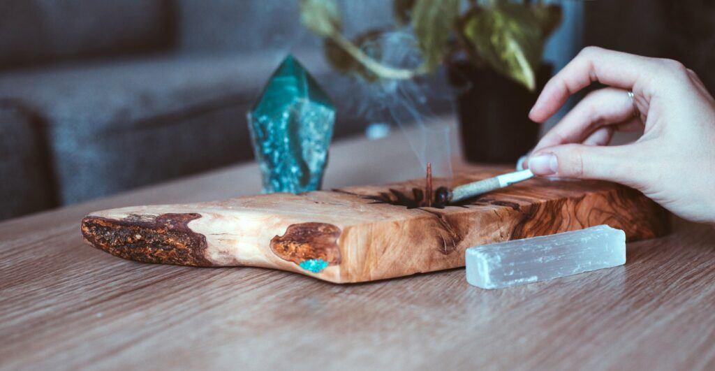 A white woman's hand ashes a pre-roll hemp joint into a handmade wooden ashtray, decorated with crystals.