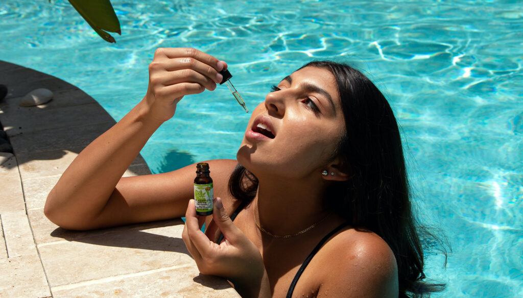 A dark-haired woman in a swimming pool takes Sir Hemp Co CBD, using the dropper top.
