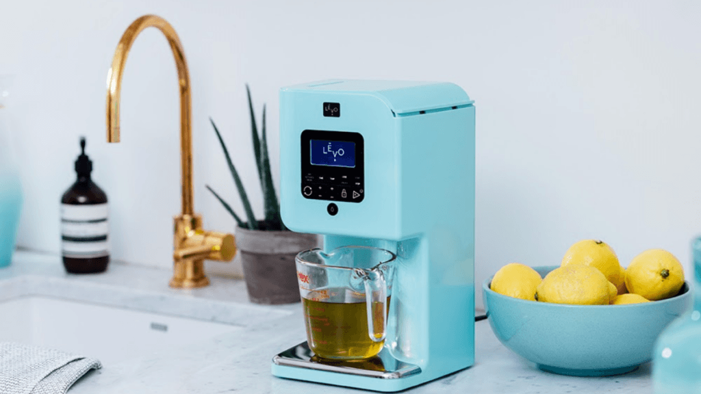 A blue Levo II Infusion Machine dispenses oil into a pyrex measuring cup. The appliance is sitting on a counter near a potted plant, a sink and a bowl of fresh lemons.