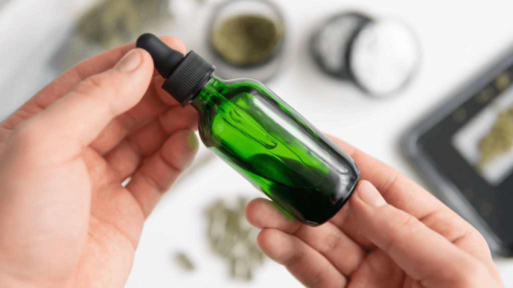A white person's hands holds a green dropper bottle of hemp extract, with hemp and smoking accessories like grinders in the background.