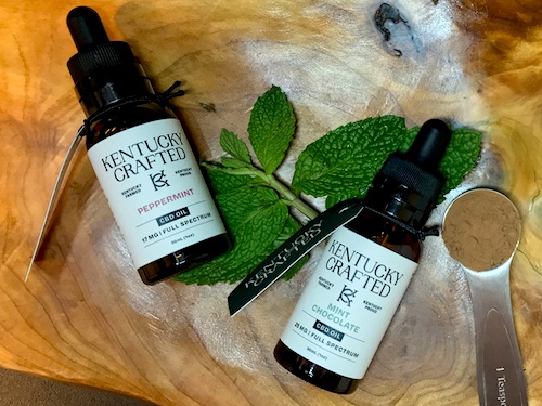 Two Kentucky Crafted CBD oil tinctures, including chcocolate mint flavor, posed with mint leaves and a spoonful of cocoa powder.