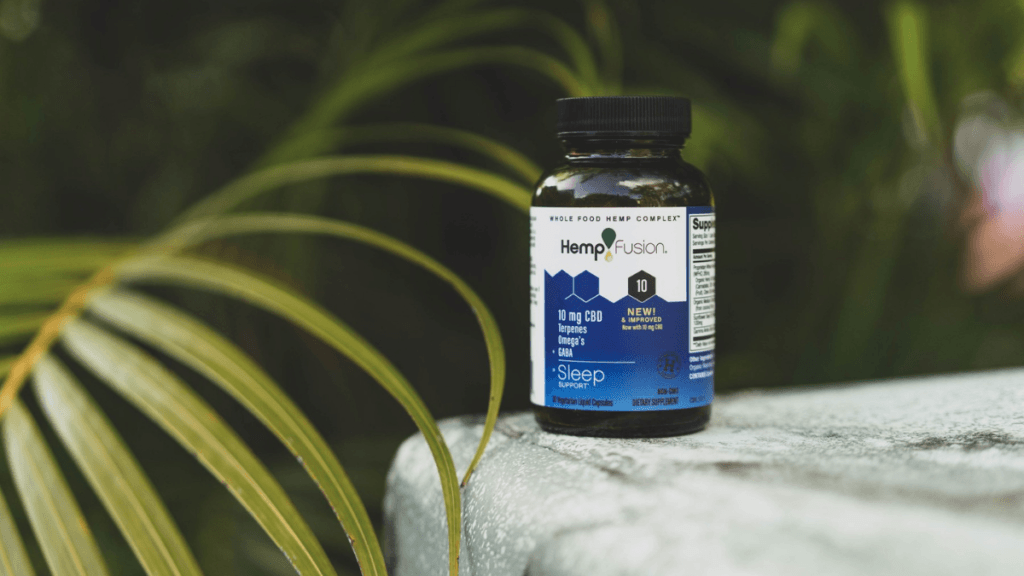 A bottle of HempFusion CBD Sleep Support capsules on a stone, surrounded by green tropical plants.