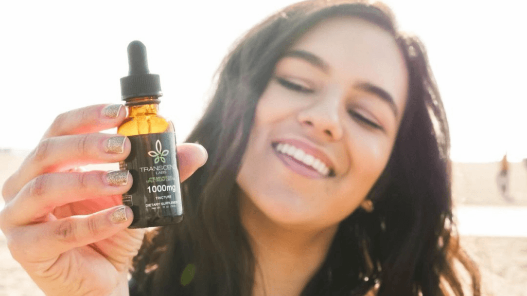 A brown-haired woman smiles as she offers a bottle of Transcend Labs Full-Spectrum CBD at the beach.