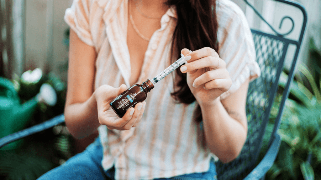 A seated person uses the Green Roads measuring wand, similar to what you would use to give a child liquid medicine, to measure out a dose of CBD oil. They are seated in an outdoor garden setting.