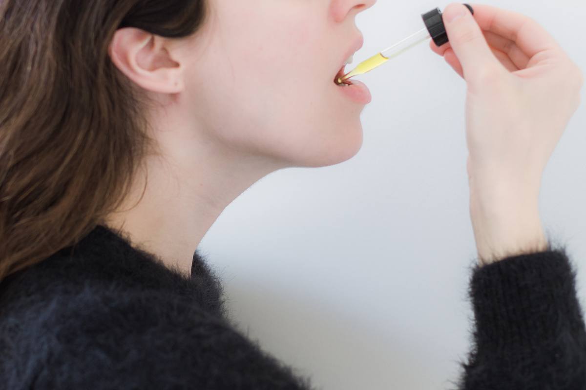 Buying CBD oil can be confusing. Use this guide to learn how to choose better CBD oil. Photo: A woman with long brown hair in a fluffy dark sweater take CBD from a dropper top.