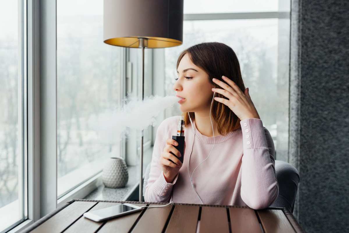 A woman seated at a slatted wooden table in the corner of a room with large windows exhales vapor while holding a vape device. Many people find the experience of vaping CBD to be extremely relaxing.