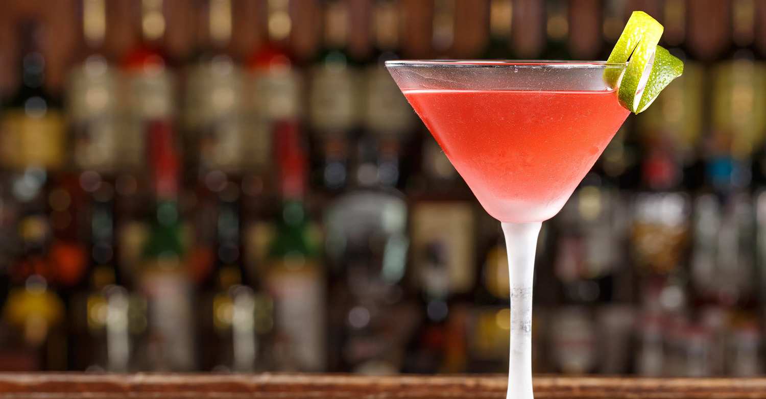 CBD cocktails are an easy way to liven up your next party. A red cocktail in a martini glass, garnished with a lemon peel, sits on a fancy bar with shelves of alcohol in the background.