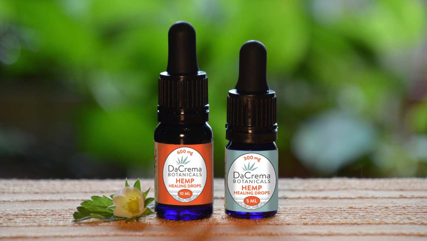 DaCrema Botanicals Hemp Healing Drops sit on a wooden counter, against a green natural background in the distance.