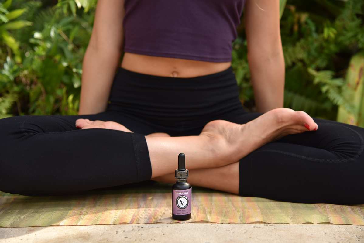 A woman sits in the lotus position on a yoga mat, with a bottle of Veritas Farms Hemp Oil Tincture at her feet.