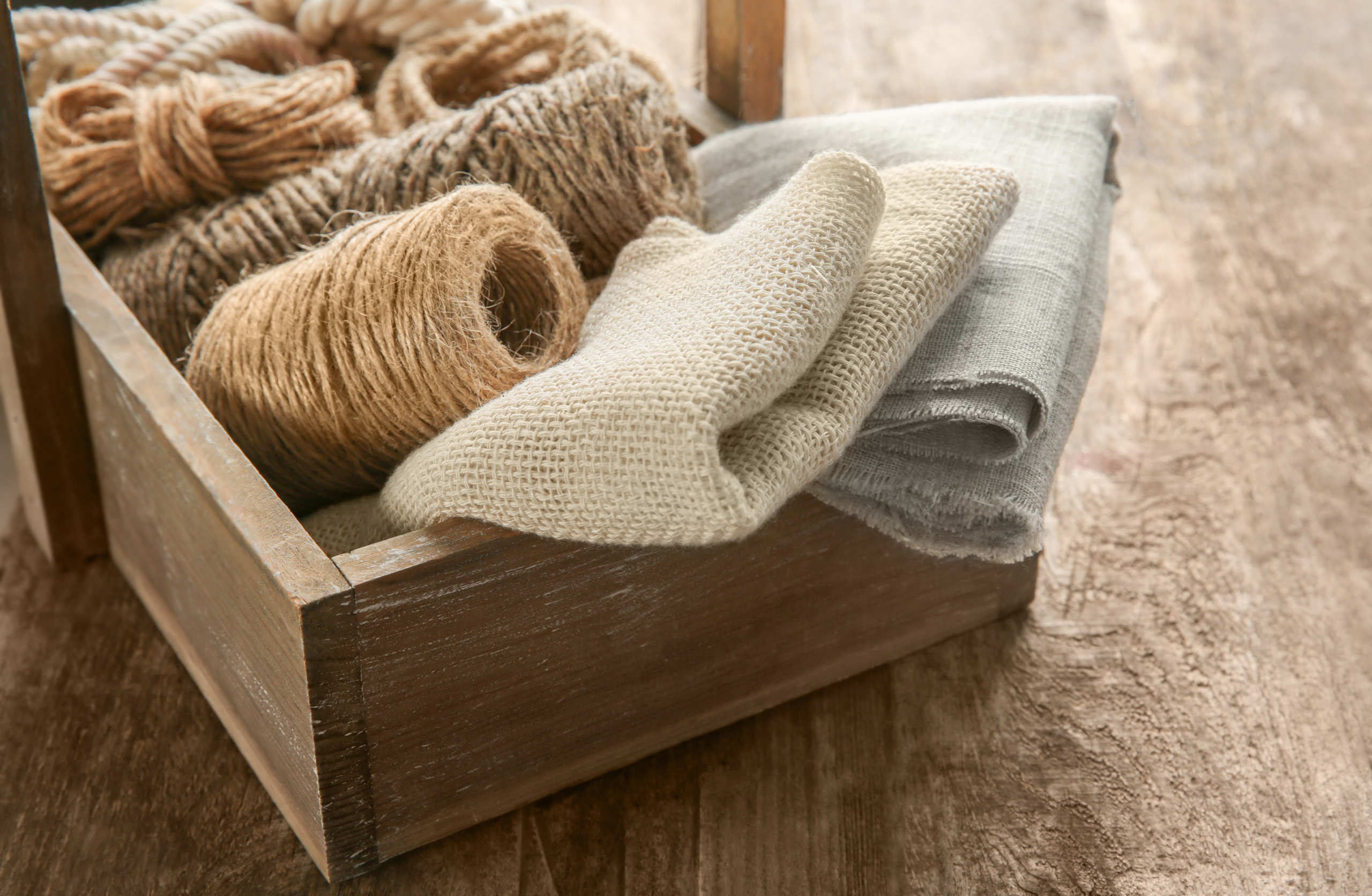 Folded hemp fabric and hemp rope and string sit in a wooden box, on a wooden table. Hemp fabric is more sustainable, stronger, and softer than other natural fabrics.