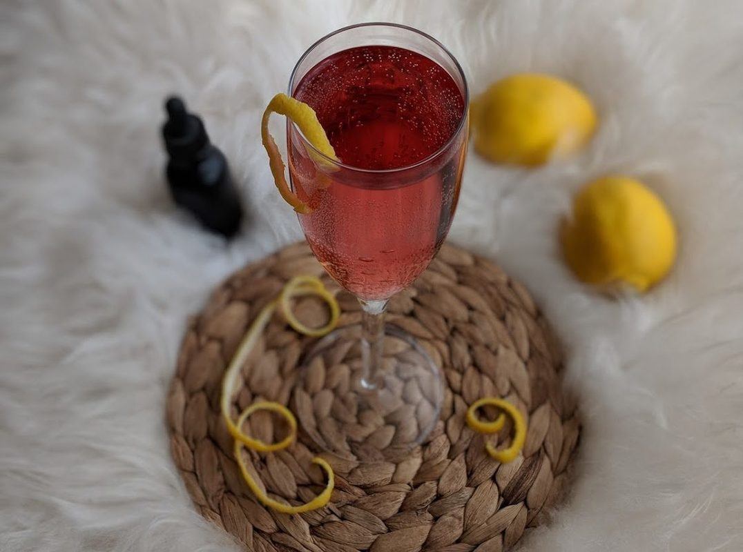 A pink cocktail in a champagne glass, garnished with a lemon twist, sits atop a woven drink coaster. Nearby are lemons and a bottle of CBD. Our sparkling CBD cocktail mixes the terpenes and flavonoids of CBD oil with sweet citrus and tart cranberry for an elevated New Years.