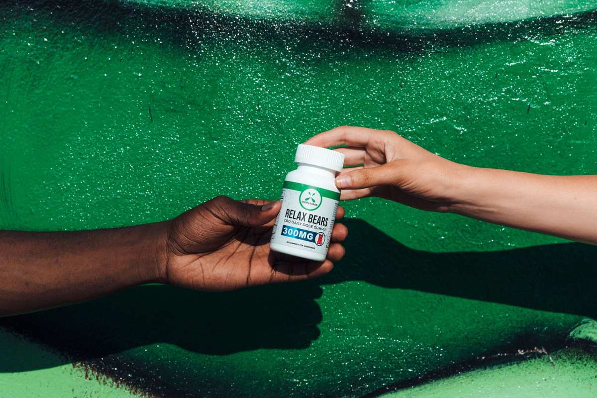 One person, seen from just the arm and hand, as they pass off a bottle of Green Roads Relax Bears to a friend, hand-to-hand. Green Roads Relax Bears combined CBD oil with a sweet taste in a convenient, chewy shape.